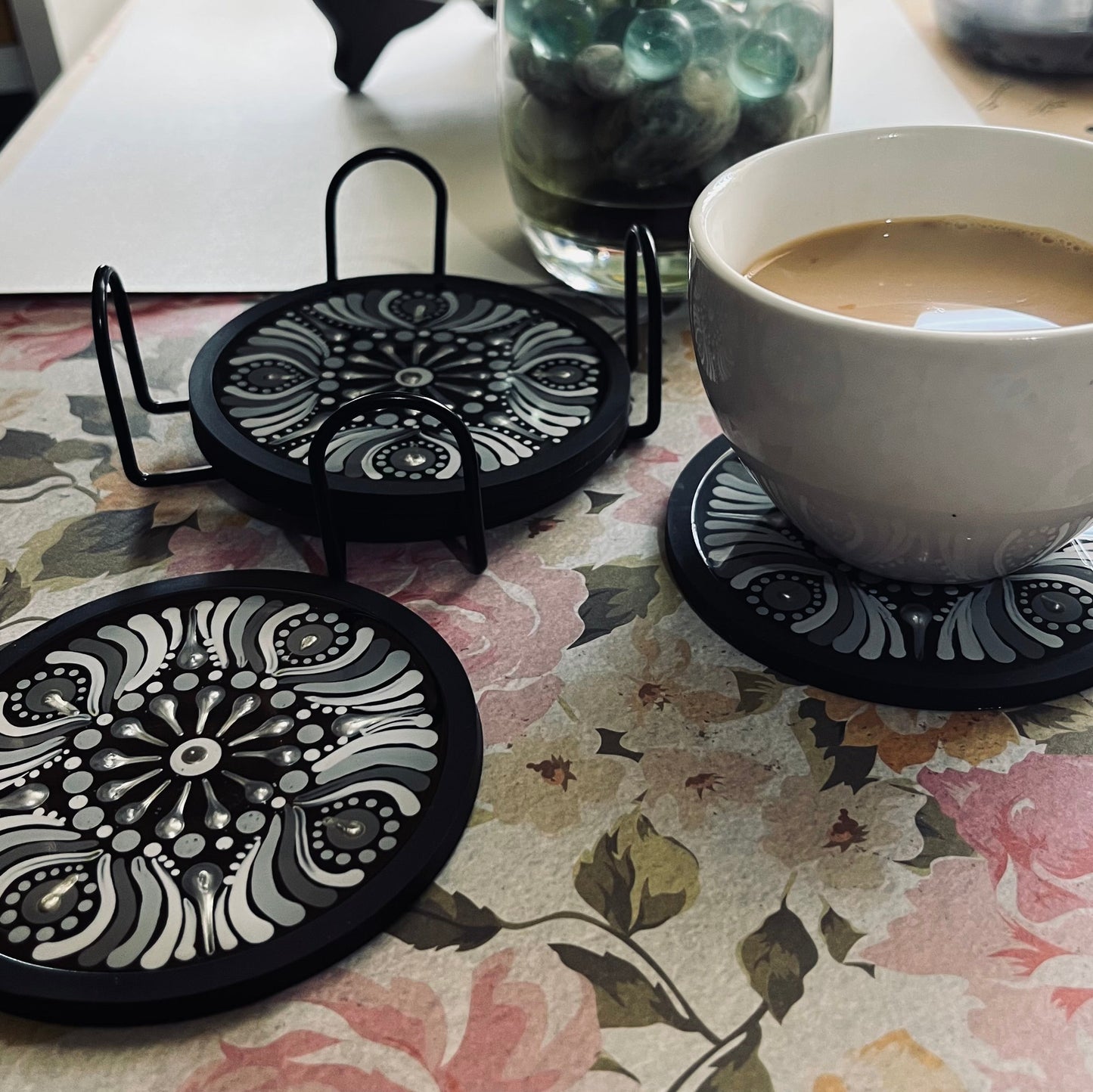 Set of black decorative coasters with a cup of coffee on a floral tablecloth.